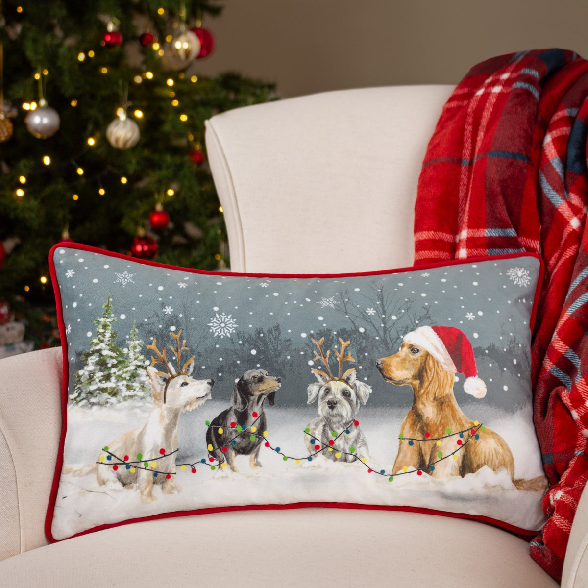 Rectangular velvet cushion with red piping, printed with four dogs in snow wearing a Santa hat and reindeer antlers, shown on an armchair by a Christmas tree.