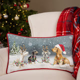 Rectangular velvet cushion with red piping, printed with four dogs in snow wearing a Santa hat and reindeer antlers, shown on an armchair by a Christmas tree.