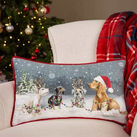 Rectangular velvet cushion with red piping, printed with four dogs in snow wearing a Santa hat and reindeer antlers, shown on an armchair by a Christmas tree.