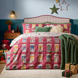 Cranberry pink Christmas Eve cotton reversible duvet cover set with illustrated townhouse windows, styled on a bed with green cushions and a throw in a festive bedroom.