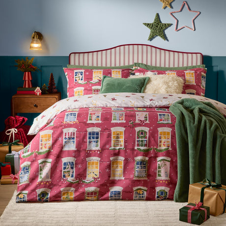 Cranberry pink Christmas Eve cotton reversible duvet cover set with illustrated townhouse windows, styled on a bed with green cushions and a throw in a festive bedroom.
