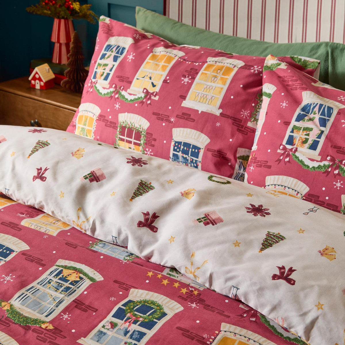 Cranberry cotton reversible duvet cover set with illustrated festive townhouse windows, shown on a bed with matching pillowcases, reversing to cream with small Christmas motifs.