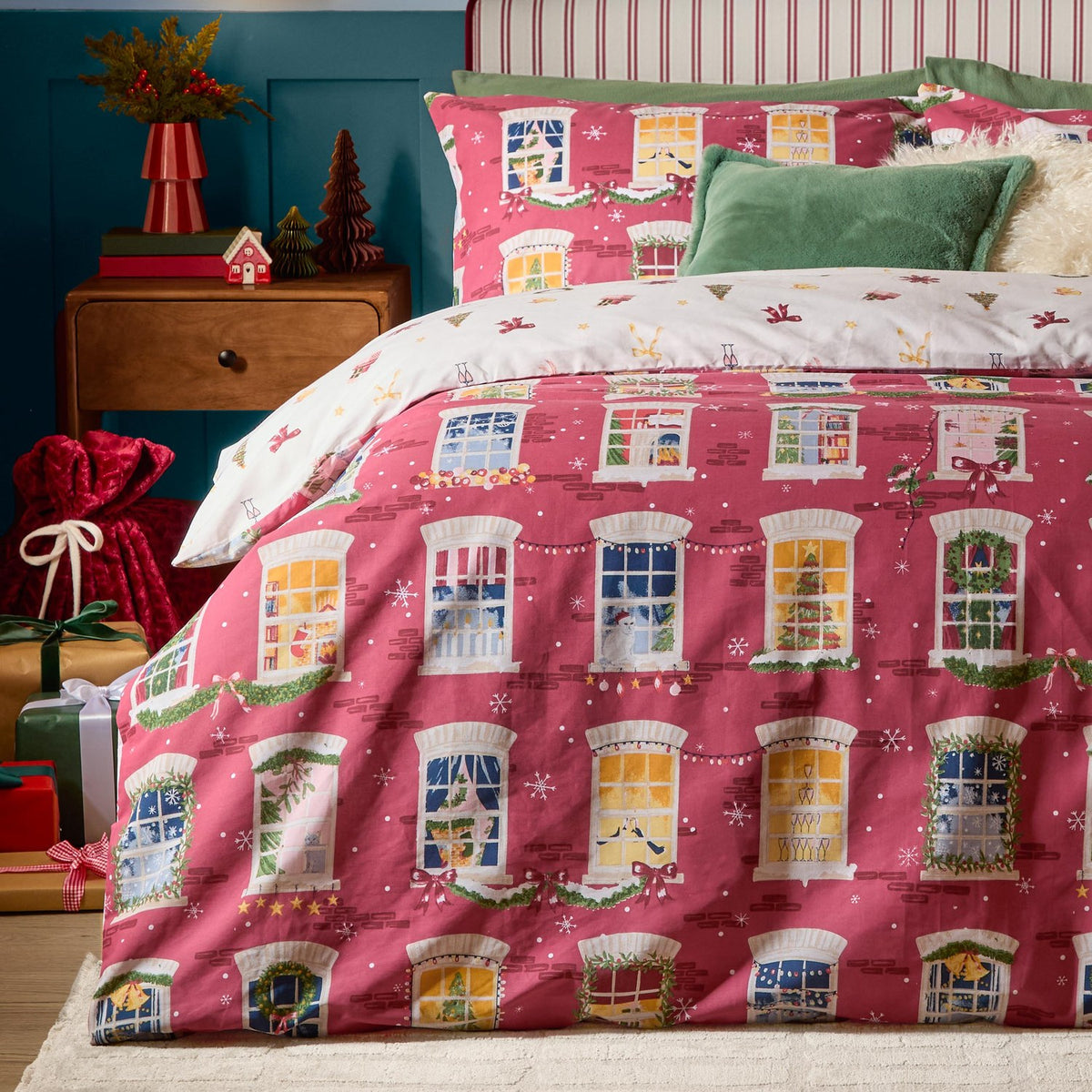 Christmas Eve Cotton Reversible Duvet Cover Set in cranberry, shown on a bedroom bed with an illustrated townhouse window and wreath print, paired with cream reverse trim and green cushions.
