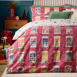 Christmas Eve Cotton Reversible Duvet Cover Set in cranberry, shown on a bedroom bed with an illustrated townhouse window and wreath print, paired with cream reverse trim and green cushions.