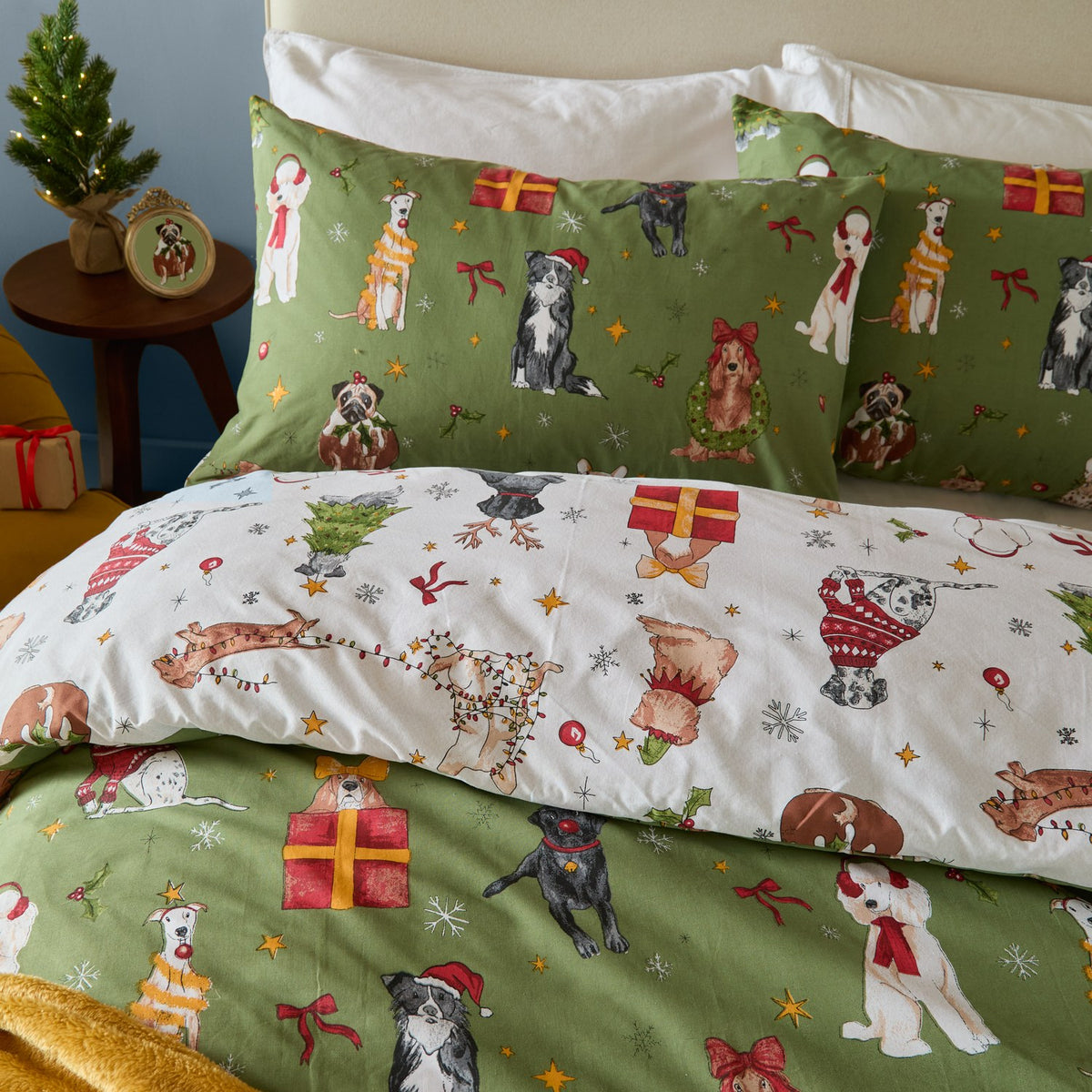 Christmas Pawty reversible cotton-blend duvet cover set on a bed, showing green side with festive dog prints and cream side with matching pattern on pillowcases.