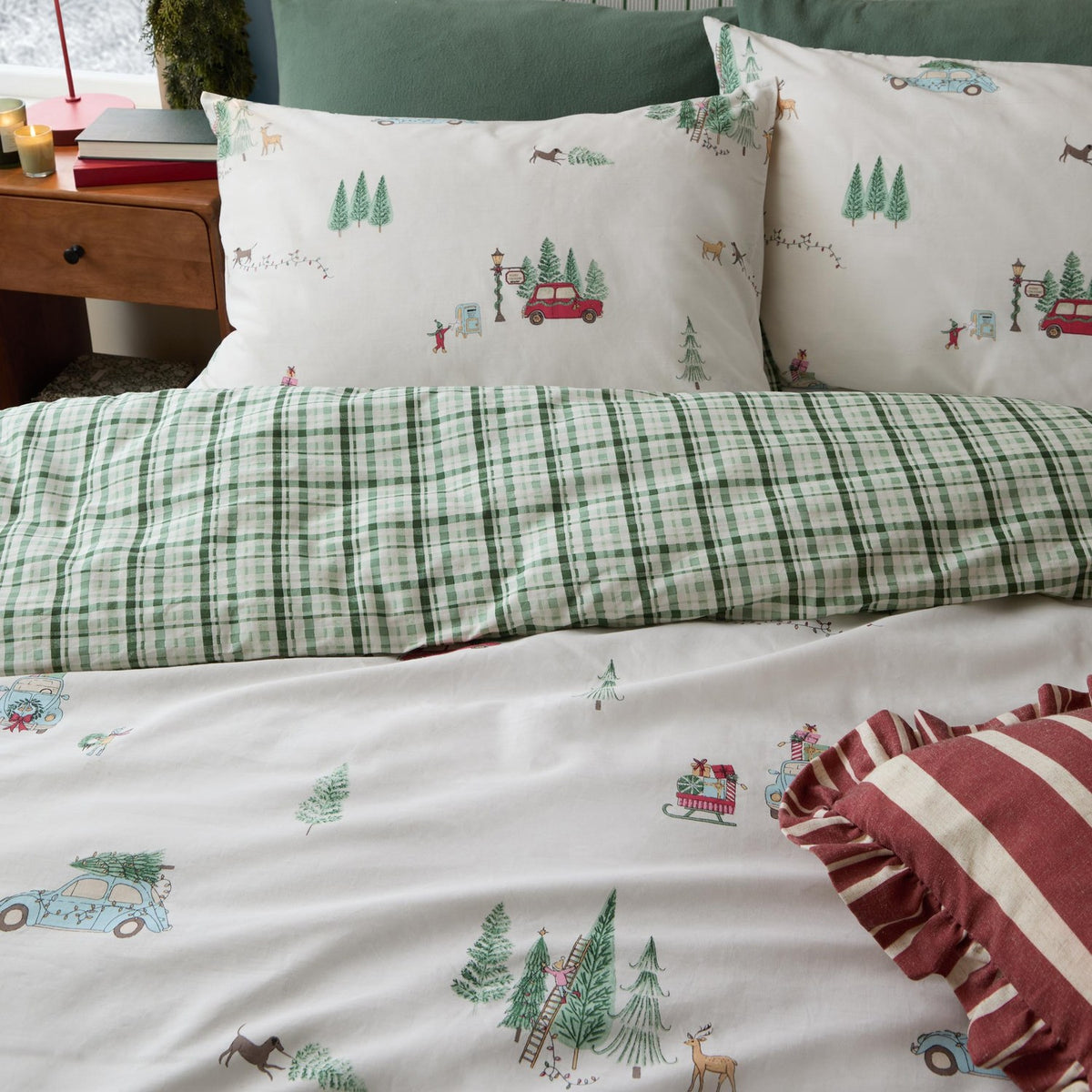 Christmas Tree Lane Duvet Cover Set in Natural/Sage, shown on a bed with pillowcases printed with cars, trees and deer, reverse side in green check, styled with a striped cushion.