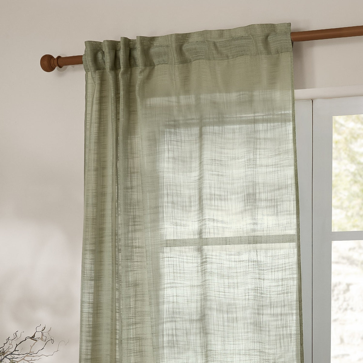 Cirrus sheer curtain panel in eucalyptus green polyester with a slubbed, cross-hatched weave, hung by hidden tab top loops on a wooden pole beside a window.
