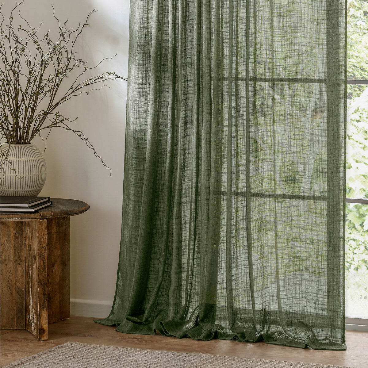 Cirrus Plain Polyester Curtain Panel in forest green, a sheer curtain with an open, slubbed weave hanging by a window beside a wooden side table and vase.