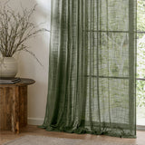 Cirrus Plain Polyester Curtain Panel in forest green, a sheer curtain with an open, slubbed weave hanging by a window beside a wooden side table and vase.