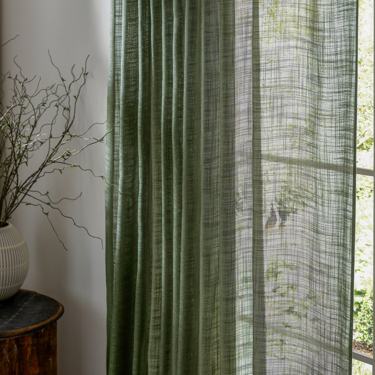 Cirrus Plain Polyester Curtain Panel in forest green, sheer pleated fabric with a slubbed woven texture, shown hanging beside a window with a vase of branches on a wooden stool.