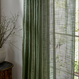 Cirrus Plain Polyester Curtain Panel in forest green, sheer pleated fabric with a slubbed woven texture, shown hanging beside a window with a vase of branches on a wooden stool.