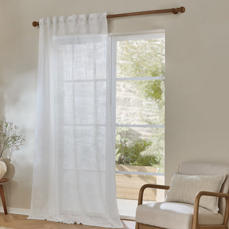 Cirrus Plain Polyester Curtain Panel in snow white sheer fabric, hung on a wooden rod beside a glass door in a living room with an armchair.