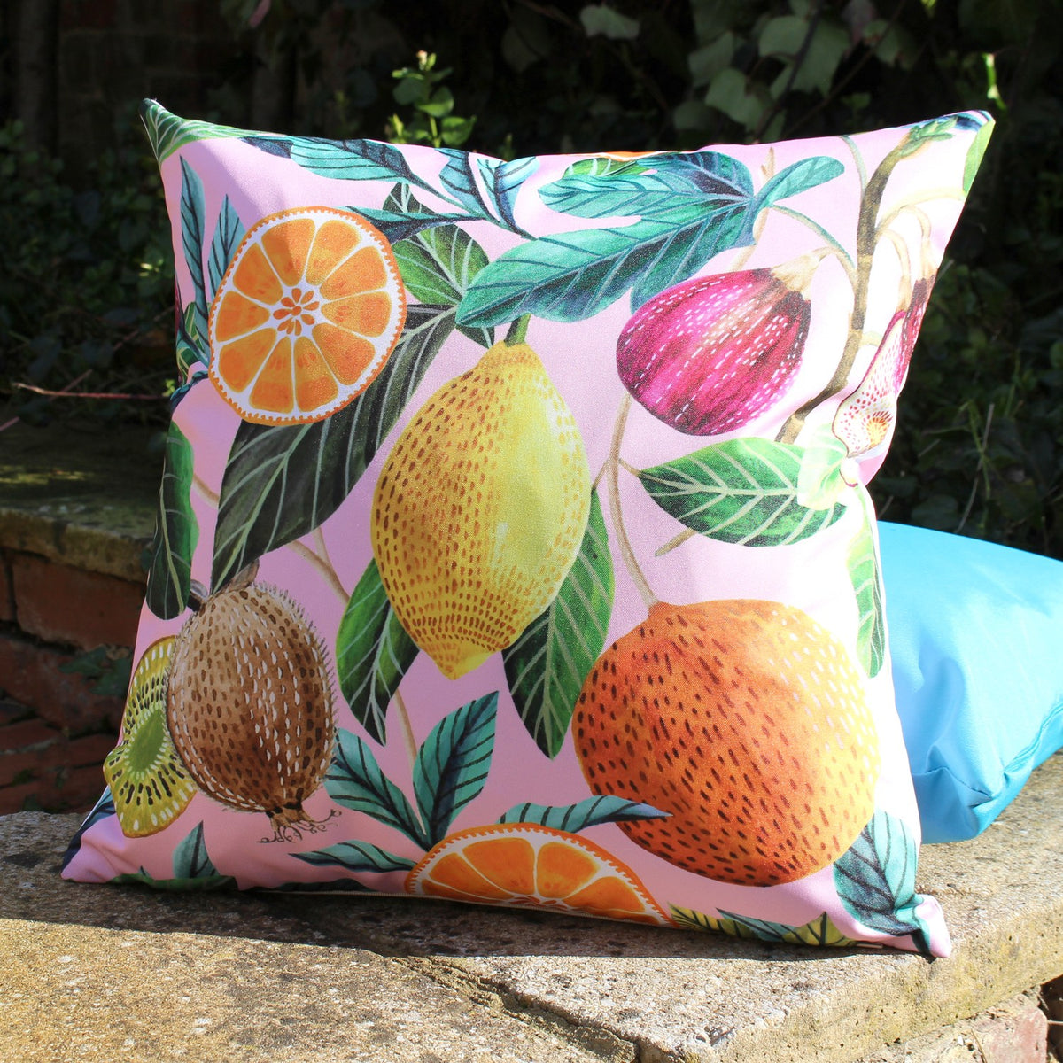 Citrus Abstract Polyester Cushion in Multi, square outdoor cushion on pink ground with watercolour print of orange slices, lemon, fig and kiwi among green leaves, shown outdoors on a stone ledge.