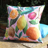 Citrus Abstract Polyester Cushion in Multi, square outdoor cushion on pink ground with watercolour print of orange slices, lemon, fig and kiwi among green leaves, shown outdoors on a stone ledge.