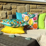 Citrus Abstract Polyester Cushion in pink with watercolour citrus fruit print, styled outdoors among teal, yellow and green cushions on a stone wall setting.