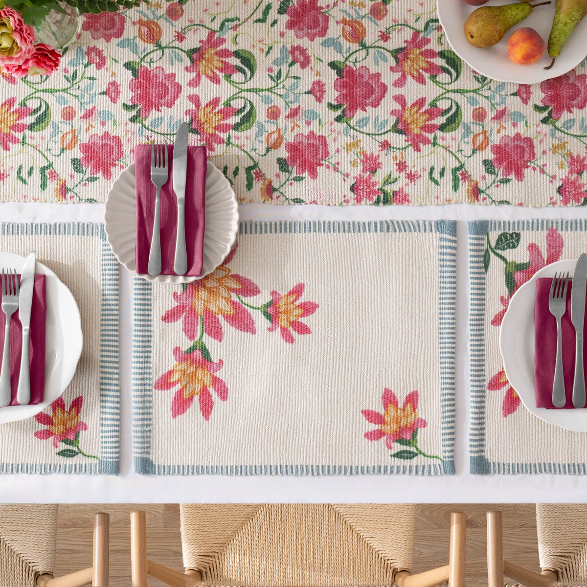 Claudia floral placemats on a dining table, cream ground with pink and amber blooms in one corner and blue-white ticking stripe border, set with white plates, cutlery, and burgundy napkins.