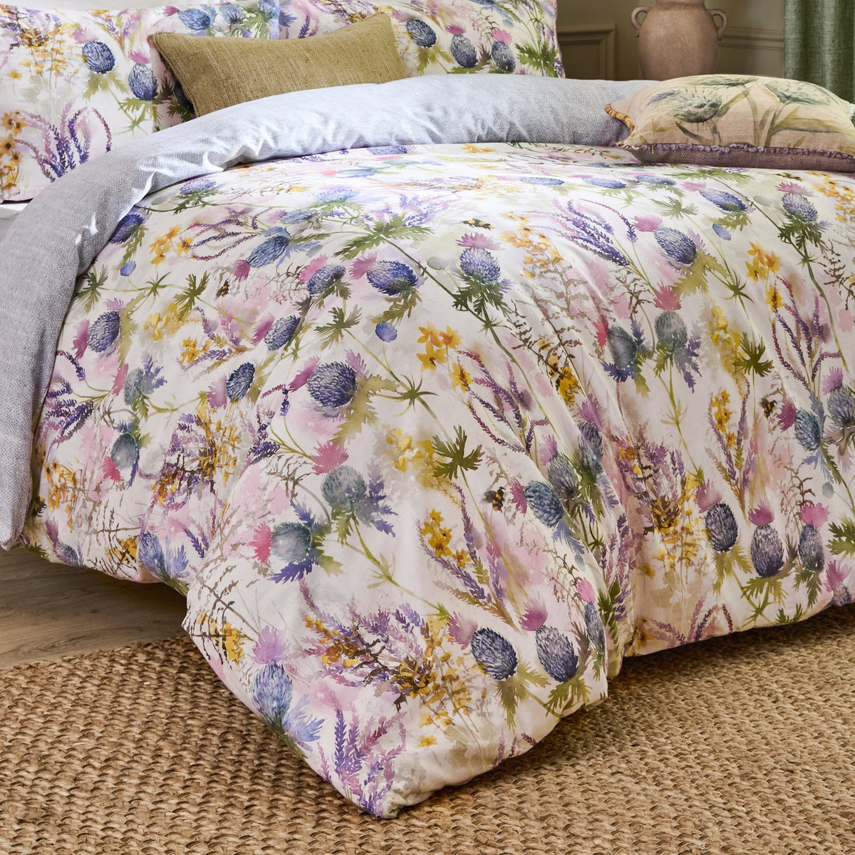 Cluaran Cotton Floral Duvet Cover Set in Multi, dressed on a bed with grey pillows, showing a cream ground print of thistles, sea holly, lavender and yellow blooms with bees, in a bedroom setting.