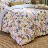 Cluaran Cotton Floral Duvet Cover Set in Multi, dressed on a bed with grey pillows, showing a cream ground print of thistles, sea holly, lavender and yellow blooms with bees, in a bedroom setting.