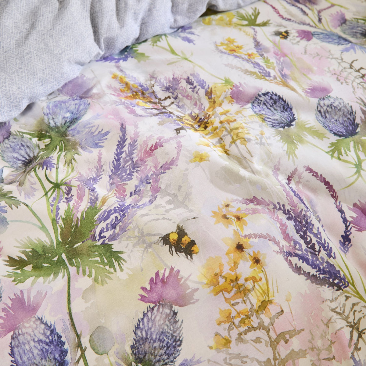 Cluaran Cotton Floral Duvet Cover Set in multi, showing a watercolour print of thistles, lavender and yellow blooms with bumblebees on a cream cotton ground, styled on a bed with grey pillows.