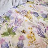 Cluaran Cotton Floral Duvet Cover Set in multi, showing a watercolour print of thistles, lavender and yellow blooms with bumblebees on a cream cotton ground, styled on a bed with grey pillows.