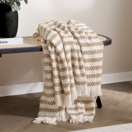 Clyne Check Polyester Throw in brown and cream, draped over a rattan bench with fringed edges, shown in a living room setting beside a black vase and books.