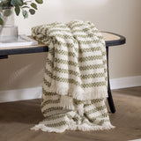 Clyne Check Polyester Throw in olive and cream woven stripes, fringed edges, draped over a black rattan-back bench beside a glass vase with foliage.