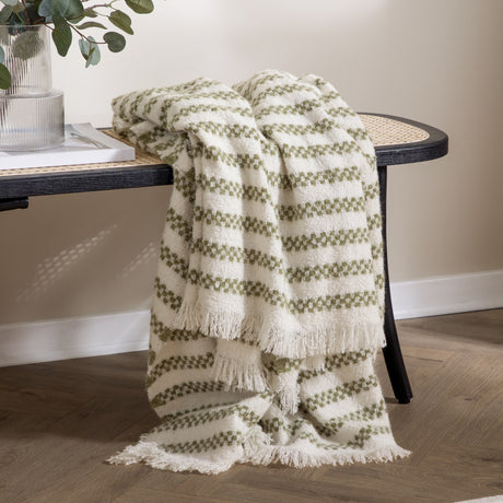Clyne Check Polyester Throw in olive and cream woven stripes, fringed edges, draped over a black rattan-back bench beside a glass vase with foliage.