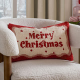 Rectangular tufted cotton cushion with "Merry Christmas" in red script on a cream ground, framed by a wavy red and pink border, shown on a cream boucle armchair.