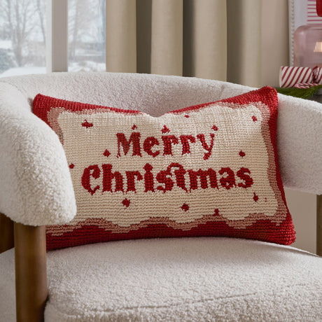 Rectangular tufted cotton cushion with "Merry Christmas" in red script on a cream ground, framed by a wavy red and pink border, shown on a cream boucle armchair.