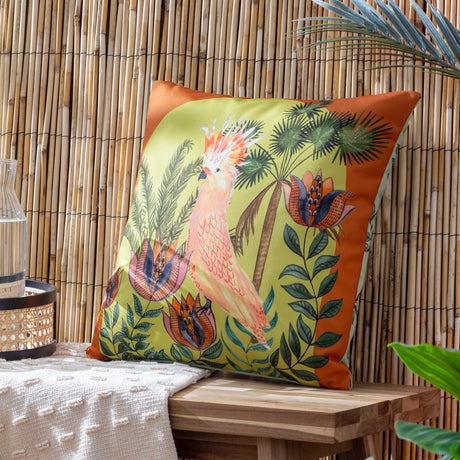 Square polyester cushion with a hand-painted cockatoo among jungle foliage on a lime-green ground with a rust orange border, shown against a bamboo backdrop.