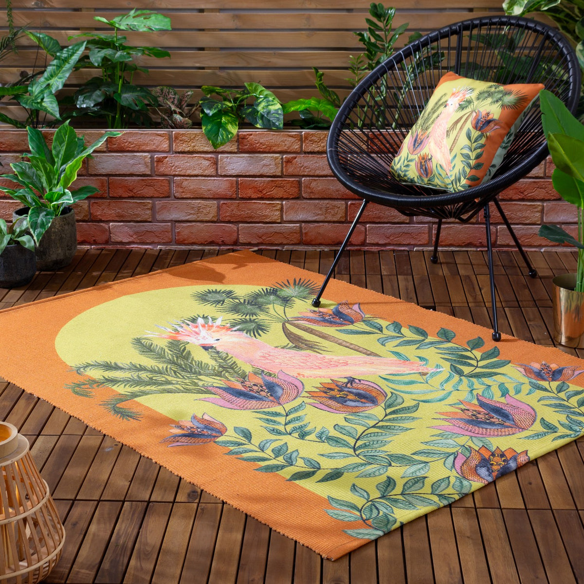 Cockatoo Reversible Polyester Cushion in multicolour with orange trim, shown on a matching patterned rug beside a black woven chair on an outdoor wooden deck.