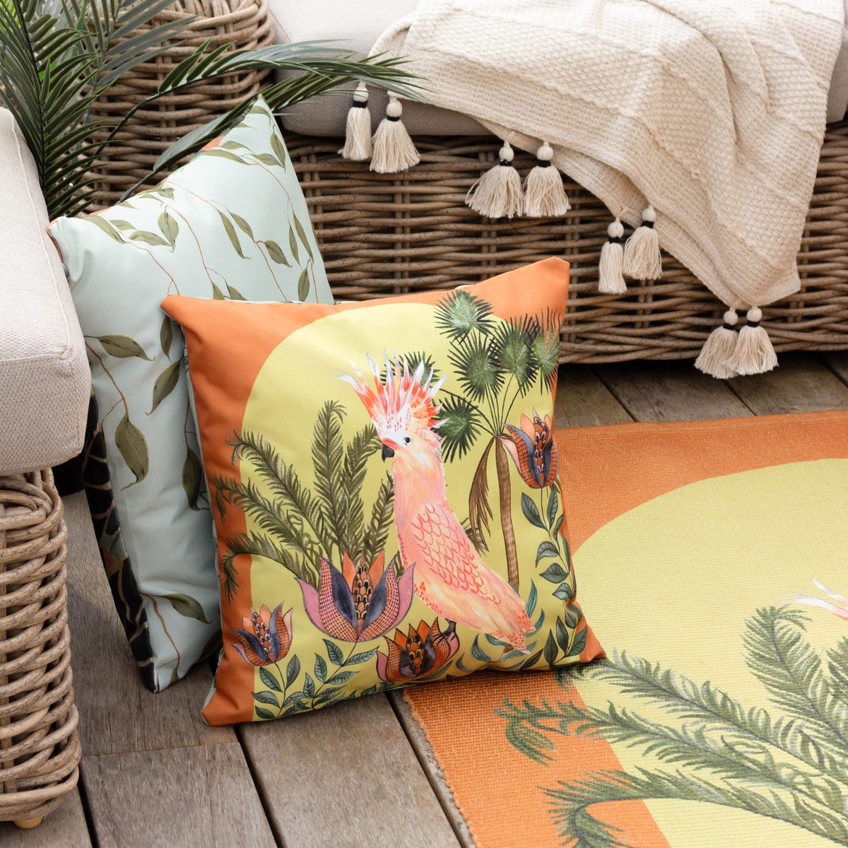 Square outdoor cushion in polyester with a hand-painted cockatoo and palm foliage design on a lime-green ground with a rust-orange border, shown on a wicker chair beside a garden rug.