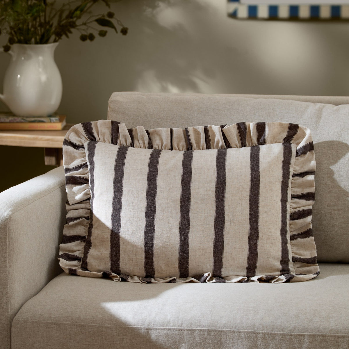 Connie Striped Linen Cushion in chocolate-brown and oatmeal, a rectangular ruffle-edged lumbar cushion resting on a beige sofa in a living room setting.
