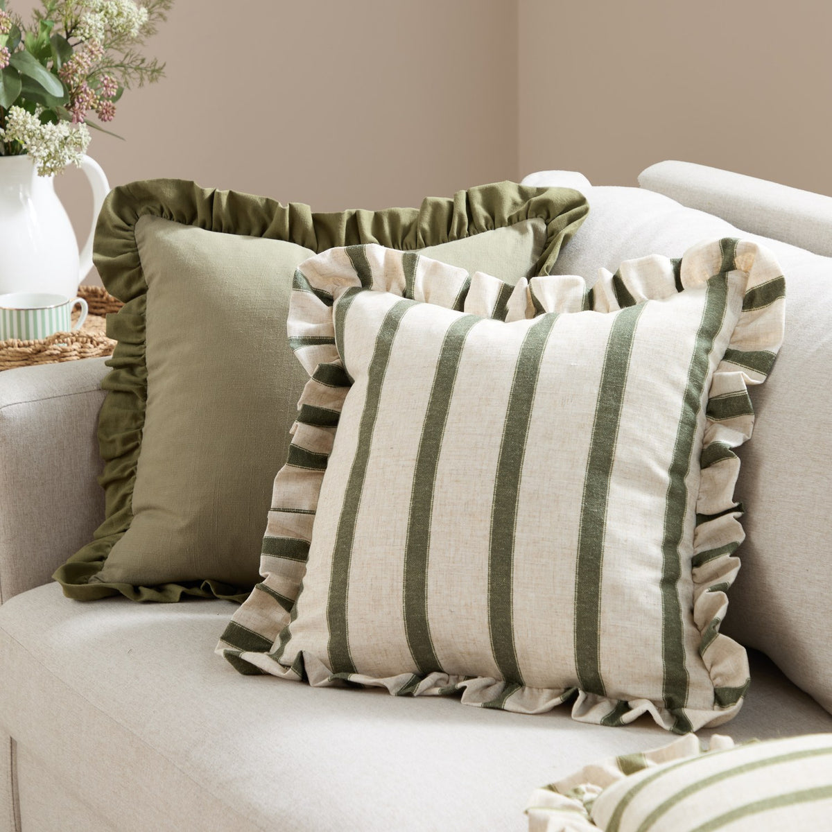 Connie Striped Linen Cushion in Moss, a square cushion with vertical moss green stripes on natural linen-look fabric and a frilled edge trim, shown on a sofa beside a plain olive ruffled cushion.