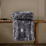 Constellation Printed Polyester Throw in charcoal and cream, folded to show a celestial eye, sun, moon and gem design with sherpa reverse, draped over a wooden bench.