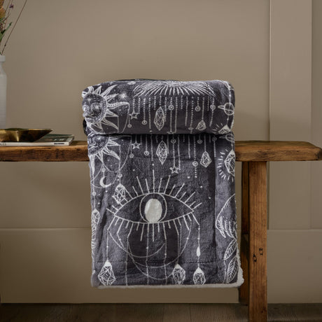 Constellation Printed Polyester Throw in charcoal and cream, folded to show a celestial eye, sun, moon and gem design with sherpa reverse, draped over a wooden bench.