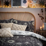 Constellation Printed Reversible Duvet Cover Set in Multi, styled on a bedroom rattan bed with charcoal celestial-print bedding, cream sun and eye motifs, and textured cushions.