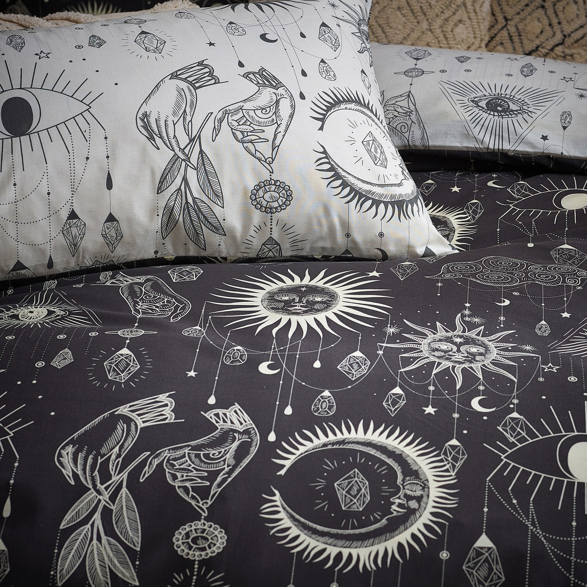 Constellation Printed Reversible Duvet Cover Set in Multi, shown on bedding with charcoal and cream sides featuring suns, moons, stars, eyes and hanging jewel motifs.
