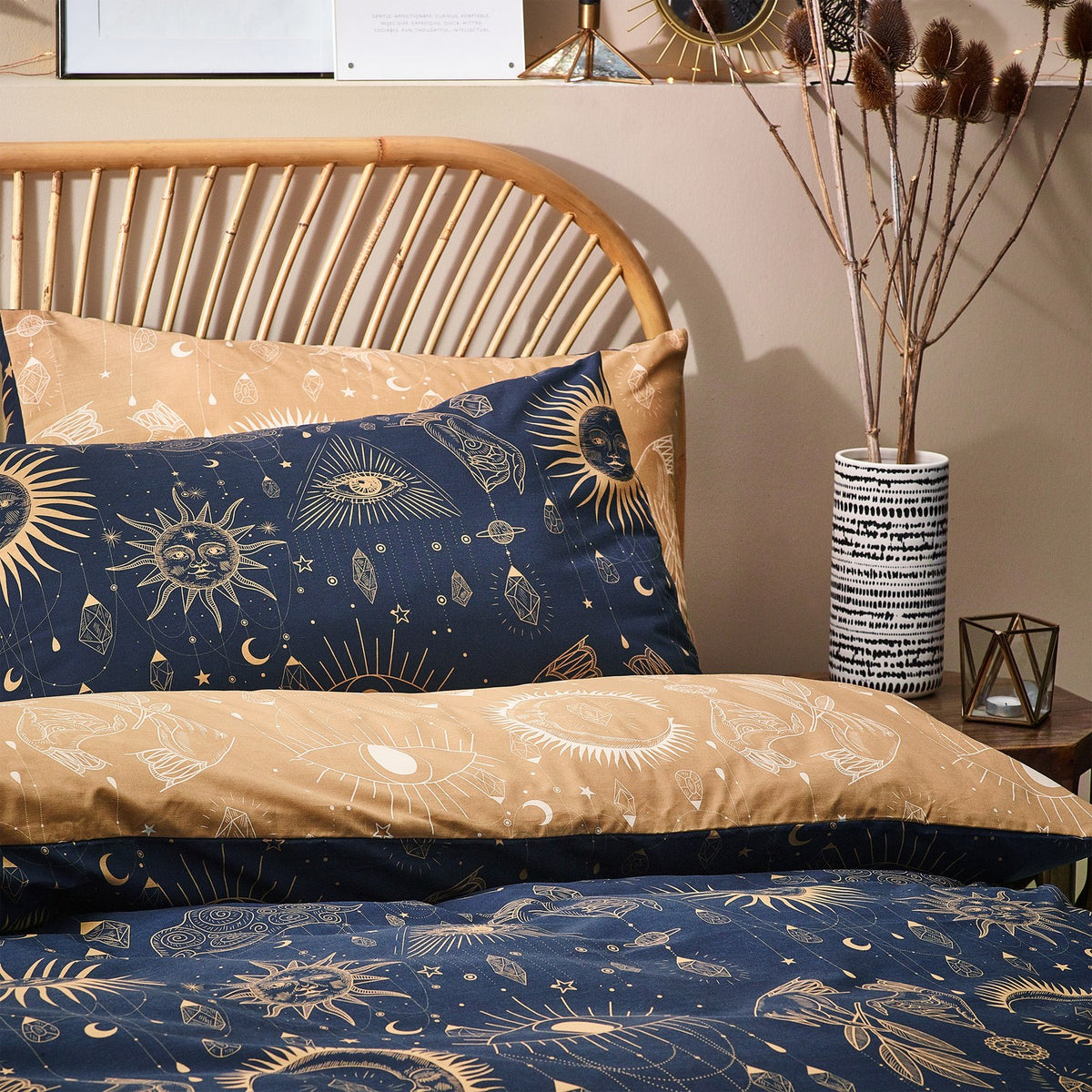 Constellation Printed Reversible Duvet Cover Set in Gold/Navy on a bed with rattan headboard, showing celestial sun, moon and star motifs on both navy and gold sides.