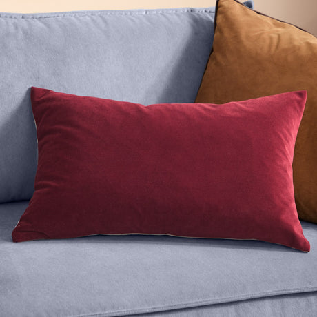 Contra Velvet Linen Cushion in oxblood, a rectangular scatter cushion with deep red velvet front, shown on a light blue sofa beside a tan velvet cushion.