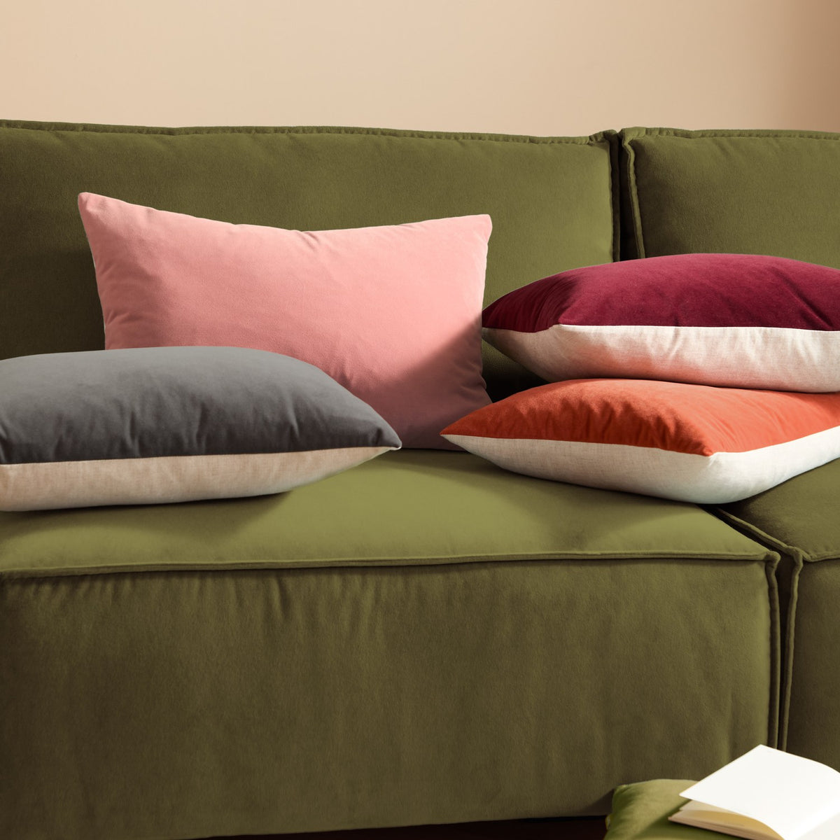 Contra Velvet Linen Cushion in oxblood, an oblong scatter cushion with deep red velvet front and neutral linen-look reverse, shown among other cushions on a green sofa.