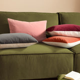 Contra Velvet Linen Cushion in oxblood, an oblong scatter cushion with deep red velvet front and neutral linen-look reverse, shown among other cushions on a green sofa.