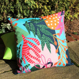 Square outdoor cushion in Coralina Abstract print, featuring tropical leaves, striped fronds and seed pods in green, coral, pink and turquoise, shown outdoors against greenery.