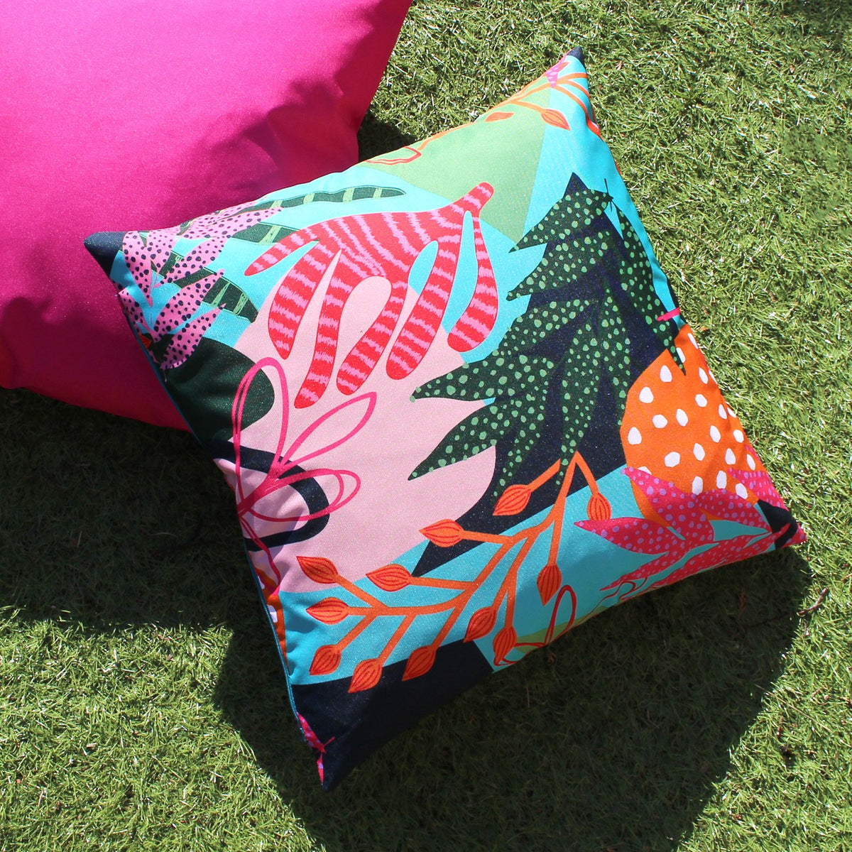 Coralina Abstract Polyester Cushion in Multi, square cover with bold tropical print in turquoise, coral, orange and navy, shown outdoors on grass beside a pink cushion.