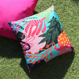 Coralina Abstract Polyester Cushion in Multi, square cover with bold tropical print in turquoise, coral, orange and navy, shown outdoors on grass beside a pink cushion.