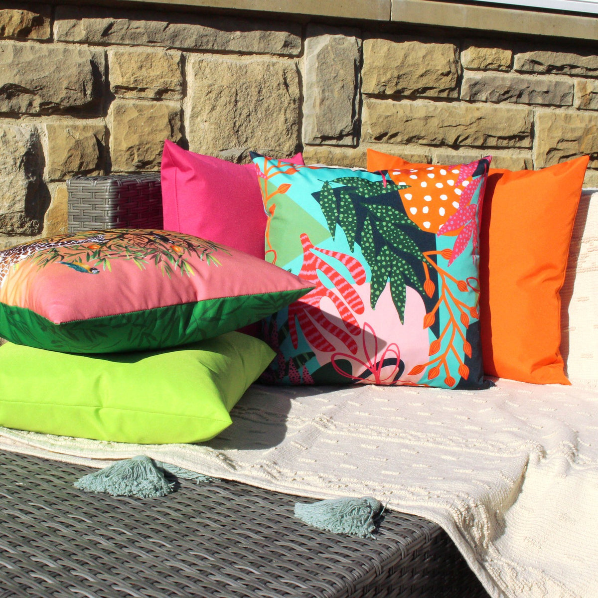 Coralina Abstract Polyester Cushion in multi tropical print with pink, orange and green cushions, styled outdoors on a rattan sofa against a stone wall.