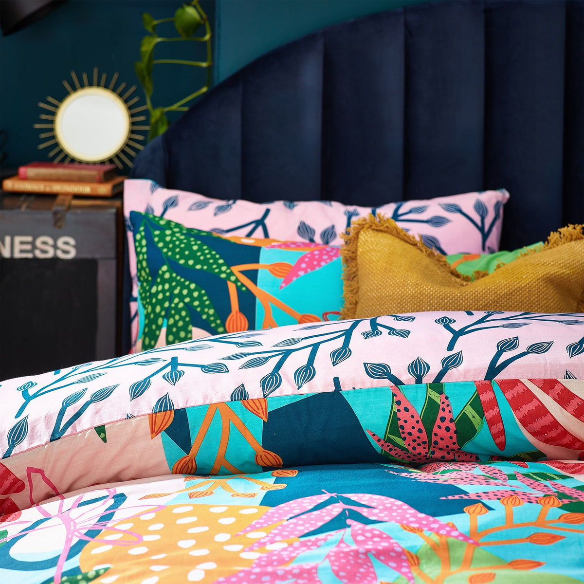 Coralina Jungle Floral Duvet Cover Set in Multi on a bed with navy headboard, showing pink reverse with navy branch print alongside the tropical multicoloured floral front and mustard fringed cushion.