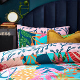 Coralina Jungle Floral Duvet Cover Set in Multi on a bed with navy headboard, showing pink reverse with navy branch print alongside the tropical multicoloured floral front and mustard fringed cushion.