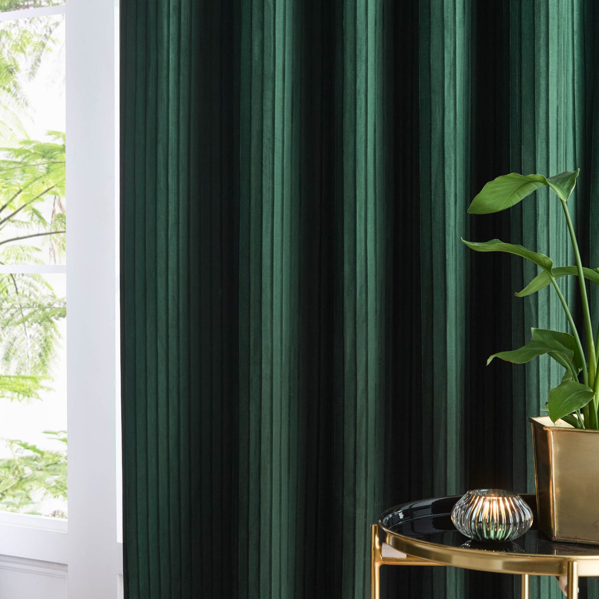Corinthian Velvet Lined Eyelet Curtains in deep green with fine vertical micro-pleat texture, shown hanging by a window beside a plant and side table.