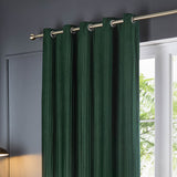 Corinthian Velvet Lined Eyelet Curtains in deep green, showing brass eyelets on a metal pole with soft micro-pleat texture, styled beside a window in a living room.